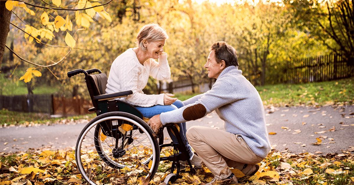 Elder woman in a wheelchair outside with her husband knelt down in front of her, and both are smiling